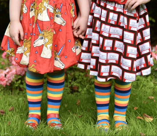 Two children wearing colorful skirts and tights standing on grass with flowers in the background wearing knee high bamboo socks